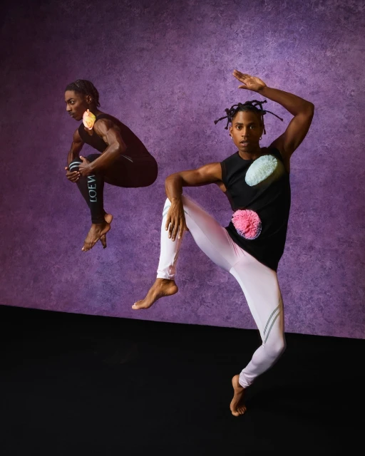 Two barefoot male dancers wearing athletic clothing with colorful pom poms pose against a purple backdrop in a studio setting.