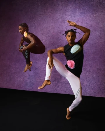 Two barefoot male dancers wearing athletic clothing with colorful pom poms pose against a purple backdrop in a studio setting.
