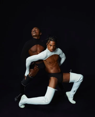 Two adult male dancers in partial bodysuits strike a pose against a dark background in a studio. Fashion and fitness concept.