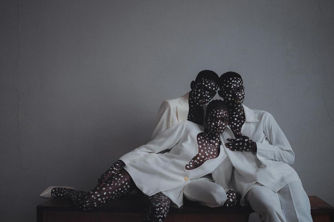 A family of three strikes a pose with white dots painted over their faces and dark tights. They are wearing matching white jackets.
