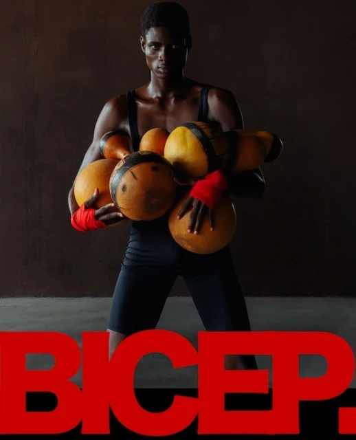 An athletic-looking Black man poses holding orange ball-shaped objects; red text 'BICEP' is in the foreground against a neutral background.