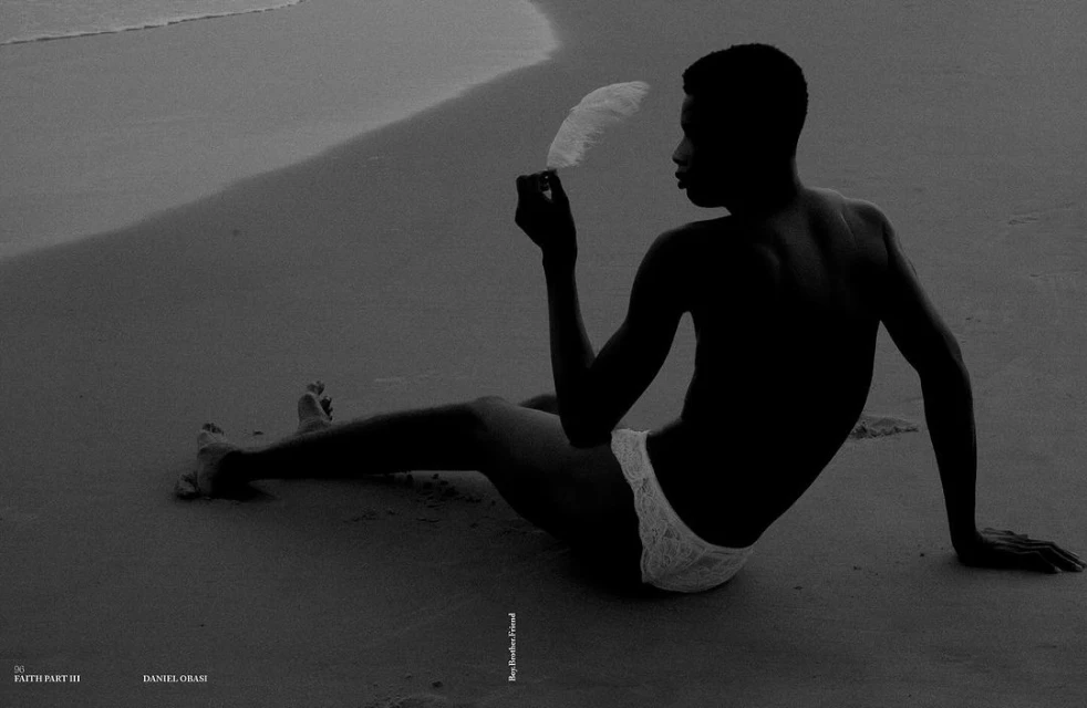 Monochromatic portrait of a shirtless man with dark skin, sitting on a sandy beach and holding a white feather near the ocean.