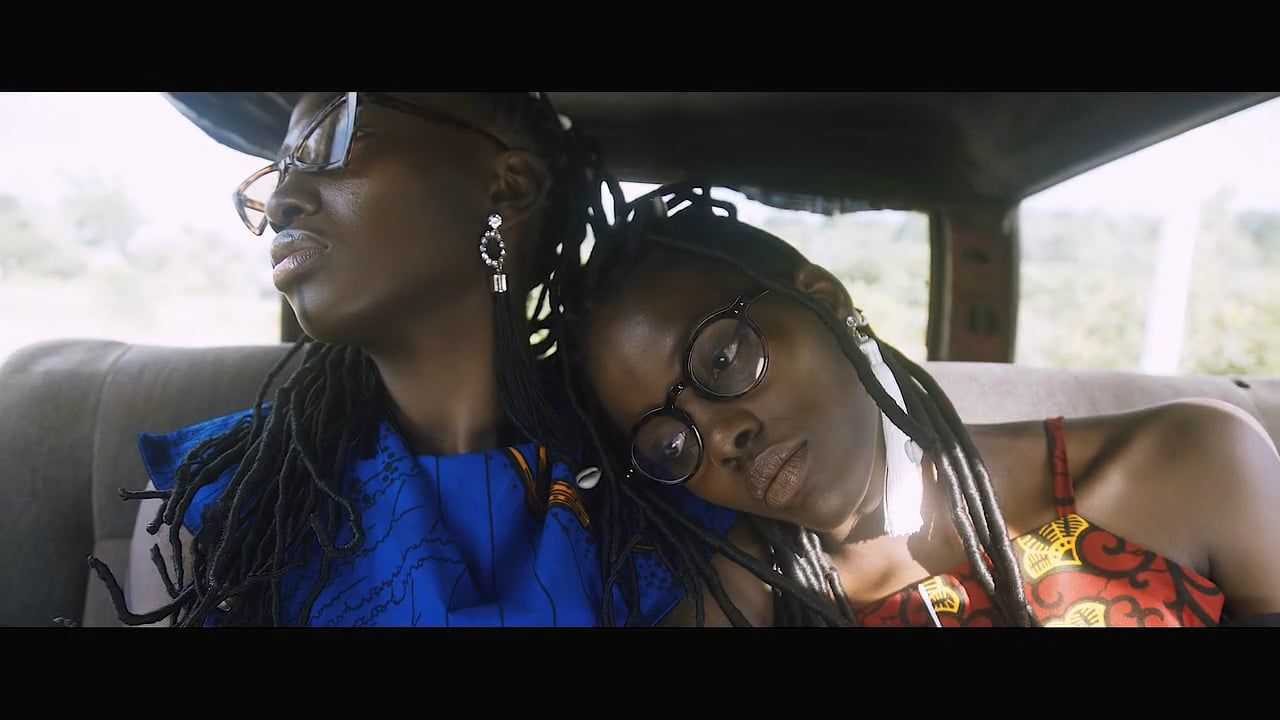 Two African women wearing glasses and traditional clothing inside a vintage car. They are resting their heads against each other.