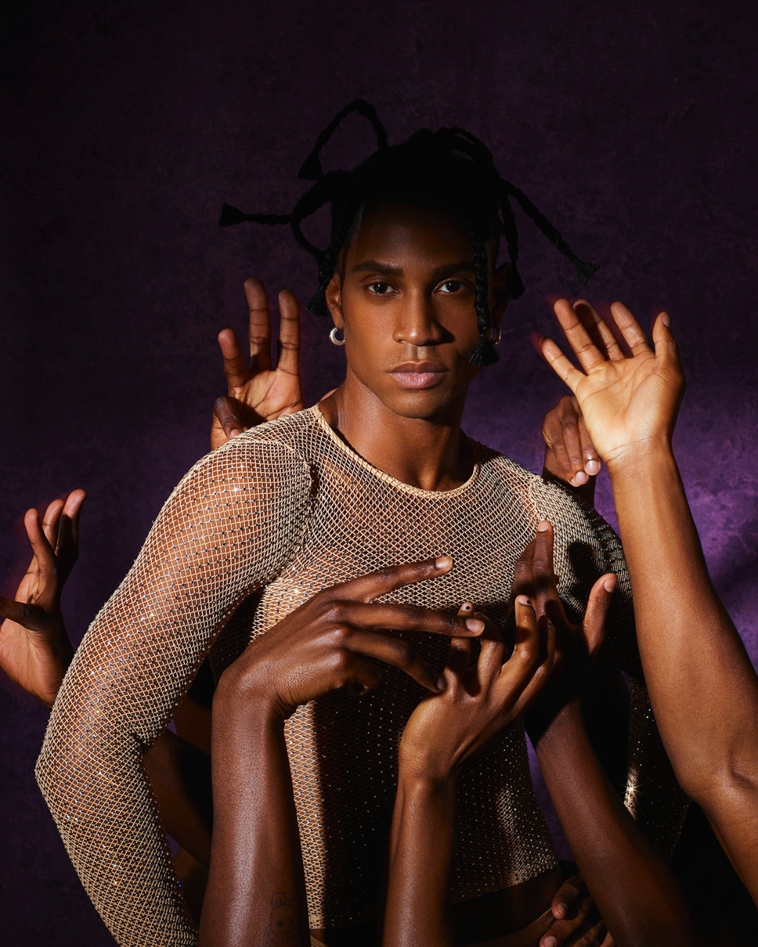 Portrait of a man with creative braids posing with multiple arms in a long sleeve top against a mottled purple background.