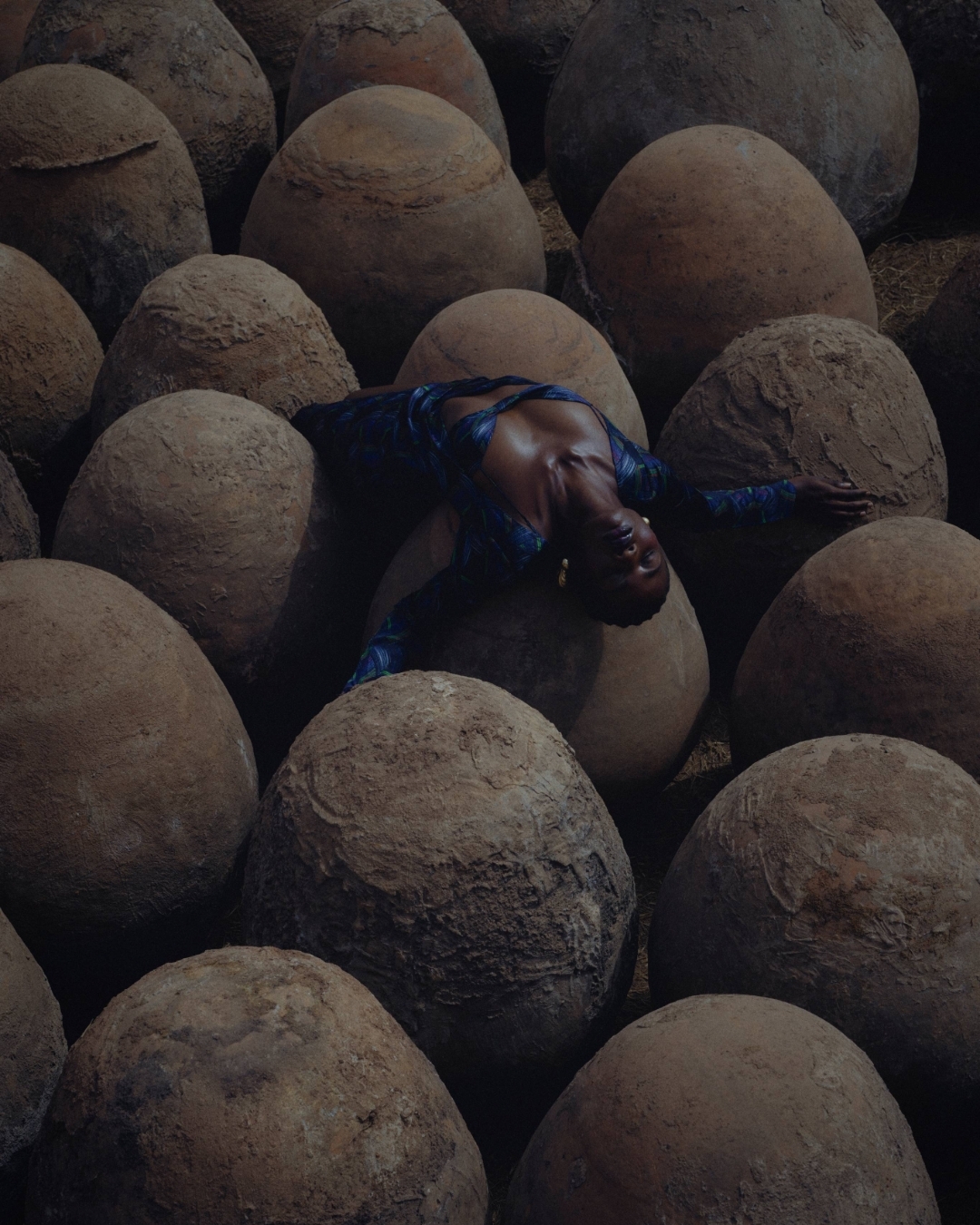 An adult female lies among many brown, spherical stones. She wears a blue patterned top and has gold earrings. Shot at eye-level.