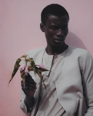 Portrait of a black man wearing a light grey jacket holding a wilted pink flower against a pink wall
