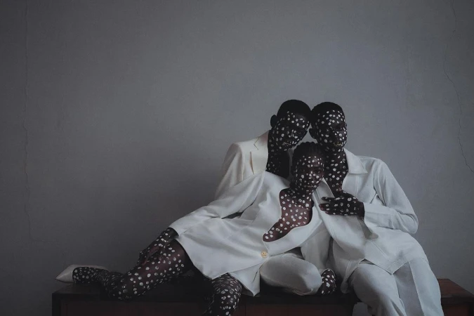 Three black models in white suits with painted faces and bodies featuring white polka dots posing together indoors.