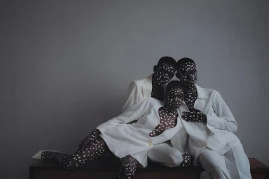 Three black models in white suits with painted faces and bodies featuring white polka dots posing together indoors.