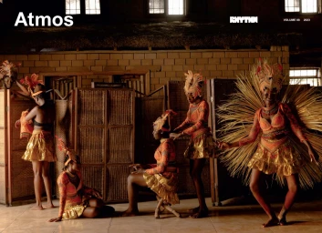 Group of black women wearing feathered headpieces and gold skirts in an interior with woven room dividers.