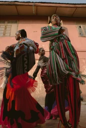 Three models wearing patterned and brightly colored garments pose against a pink building exterior