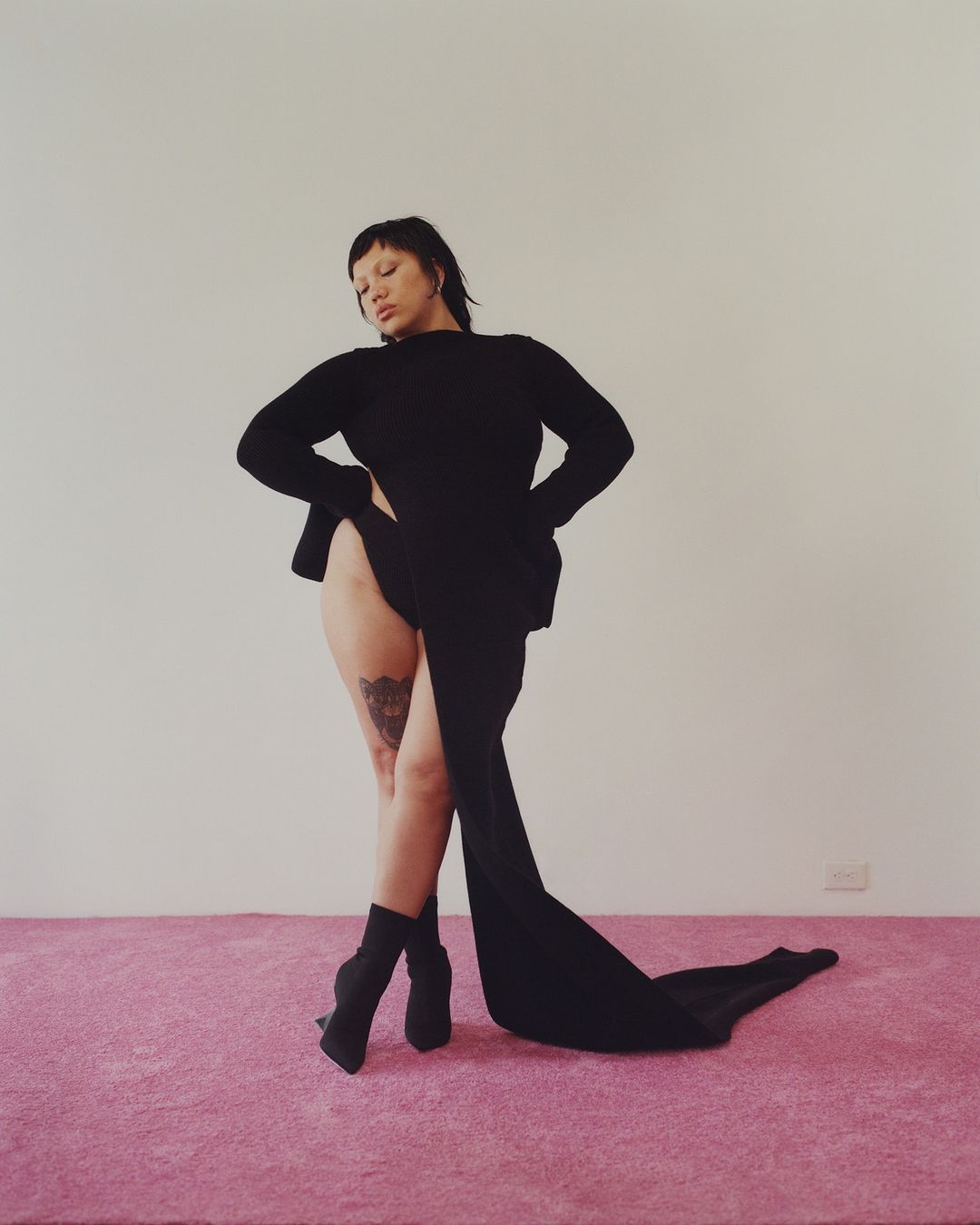 Model in black long-sleeve dress and boots standing on pink carpet against white wall photographed by Daniel Jack Lyons