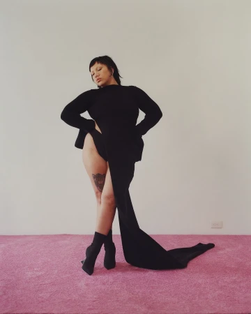 Model in black long-sleeve dress and boots standing on pink carpet against white wall photographed by Daniel Jack Lyons