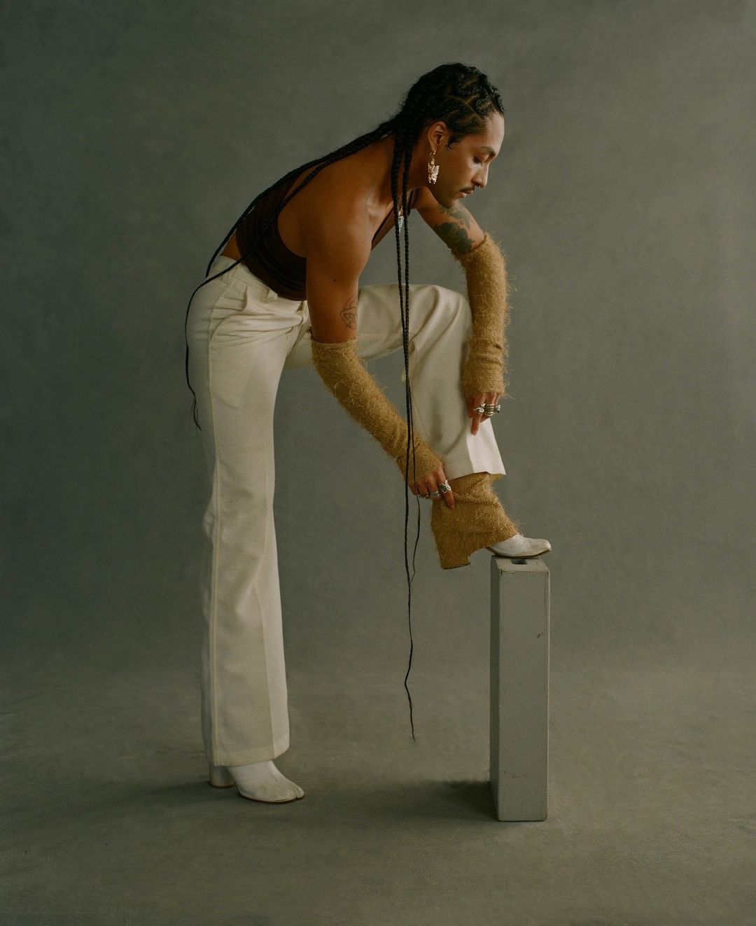 A person with braids poses in a brown tank top and white pants, placing a foot on a gray block in a studio setting.