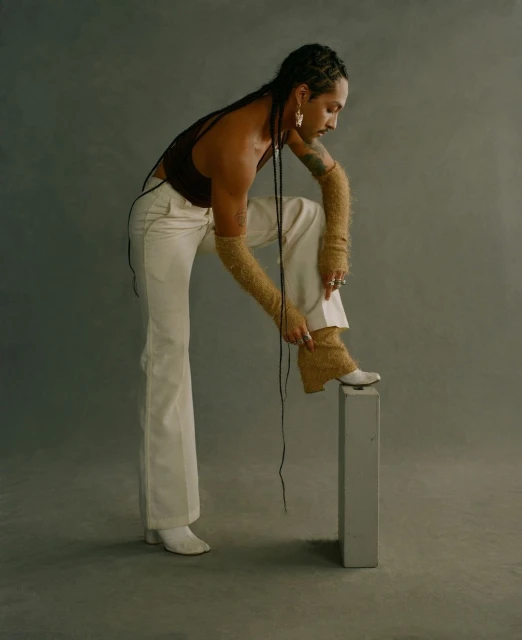 A person with braids poses in a brown tank top and white pants, placing a foot on a gray block in a studio setting.