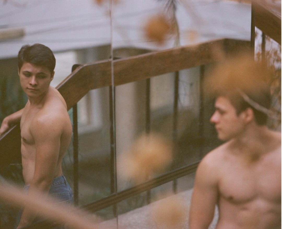 Two bare-chested men in blue jeans stand outdoors on a staircase with wooden railings, looking in different directions with serious expressions.