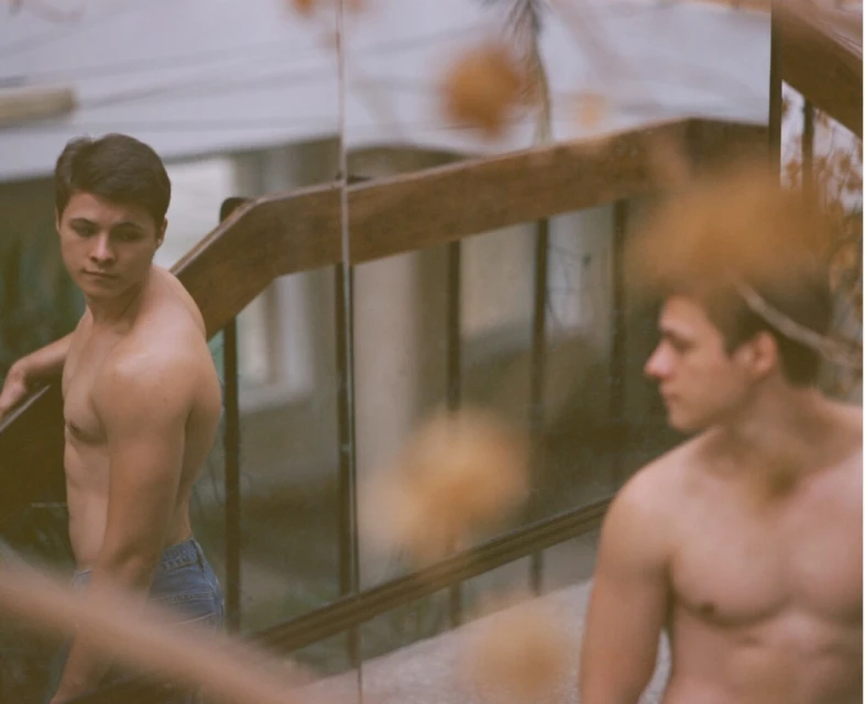 Two bare-chested men in blue jeans stand outdoors on a staircase with wooden railings, looking in different directions with serious expressions.