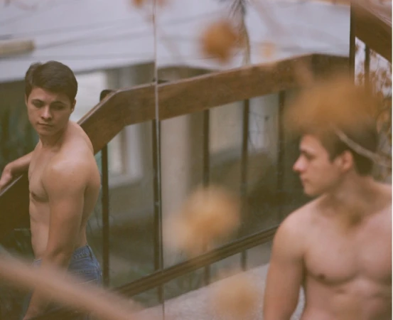 Two bare-chested men in blue jeans stand outdoors on a staircase with wooden railings, looking in different directions with serious expressions.