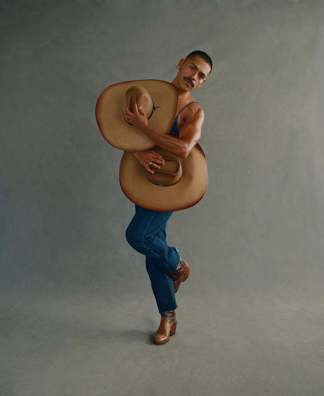Portrait of a muscular male model wearing denim and boots, posing with two cowboy hats in a photo studio against a neutral background.