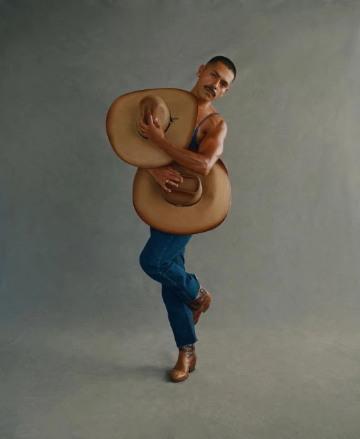 Portrait of a muscular male model wearing denim and boots, posing with two cowboy hats in a photo studio against a neutral background.