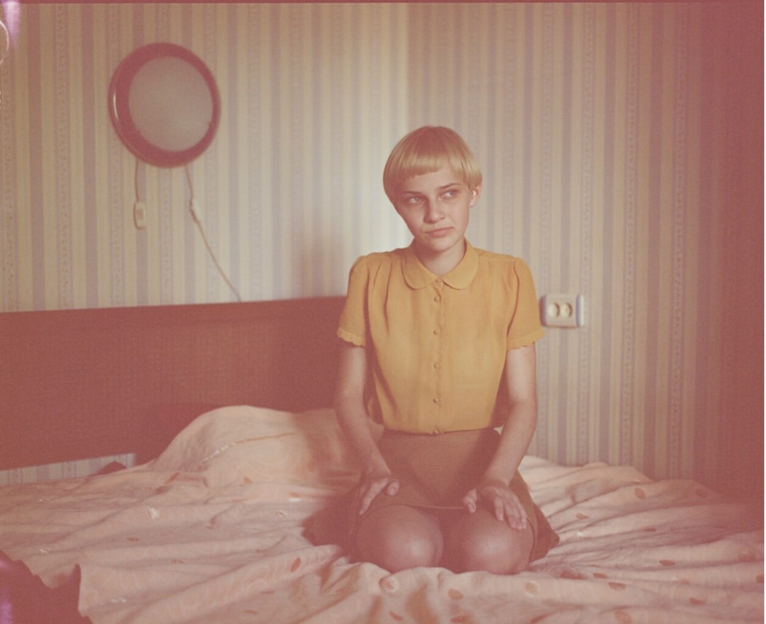 A young woman with a short blonde hairstyle sits on a bed in a bedroom setting, wearing a mustard-colored blouse and a brown skirt.