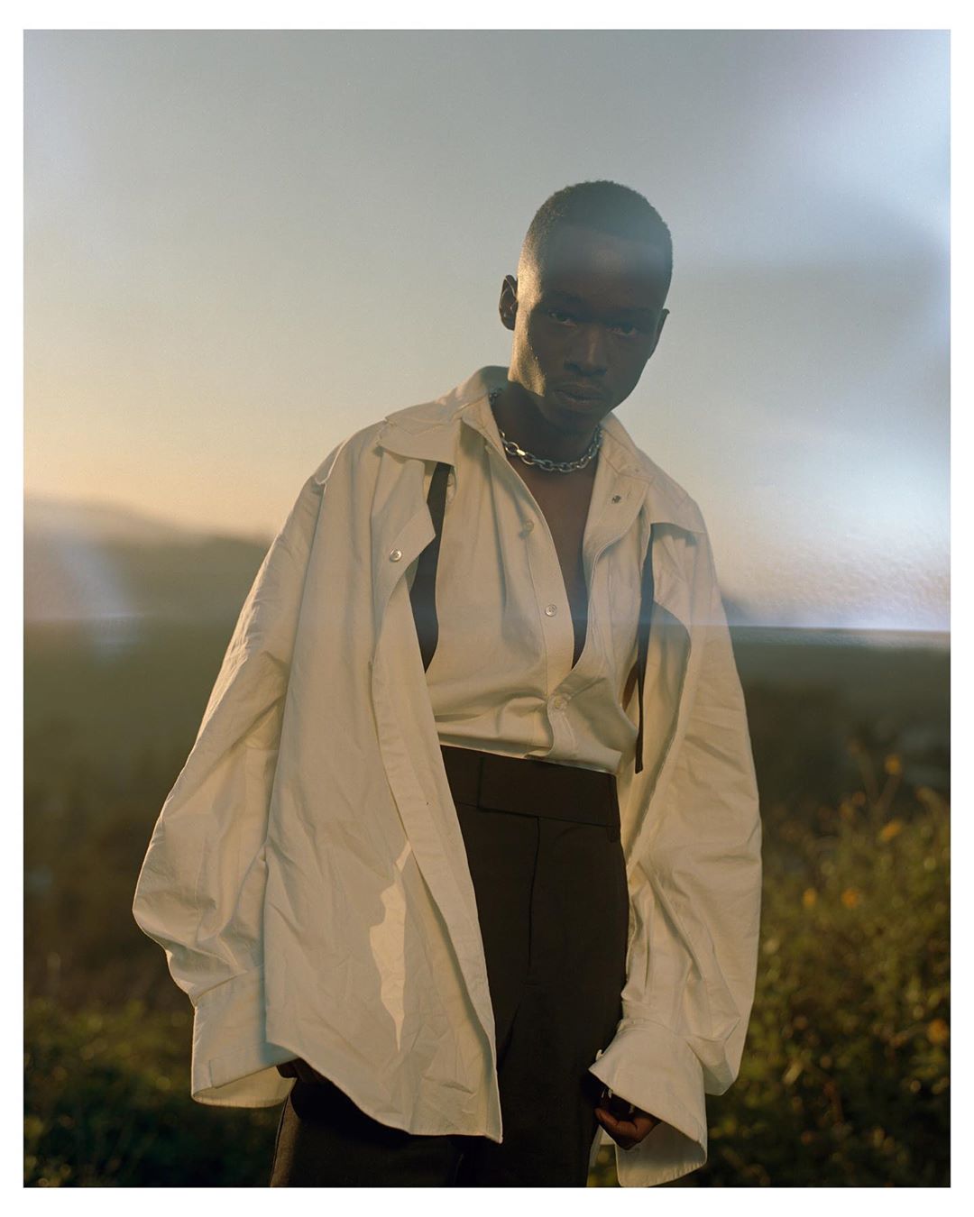 Portrait of a young Black man in a loose, unbuttoned beige shirt and dark trousers, outdoors during golden hour with soft focus background.