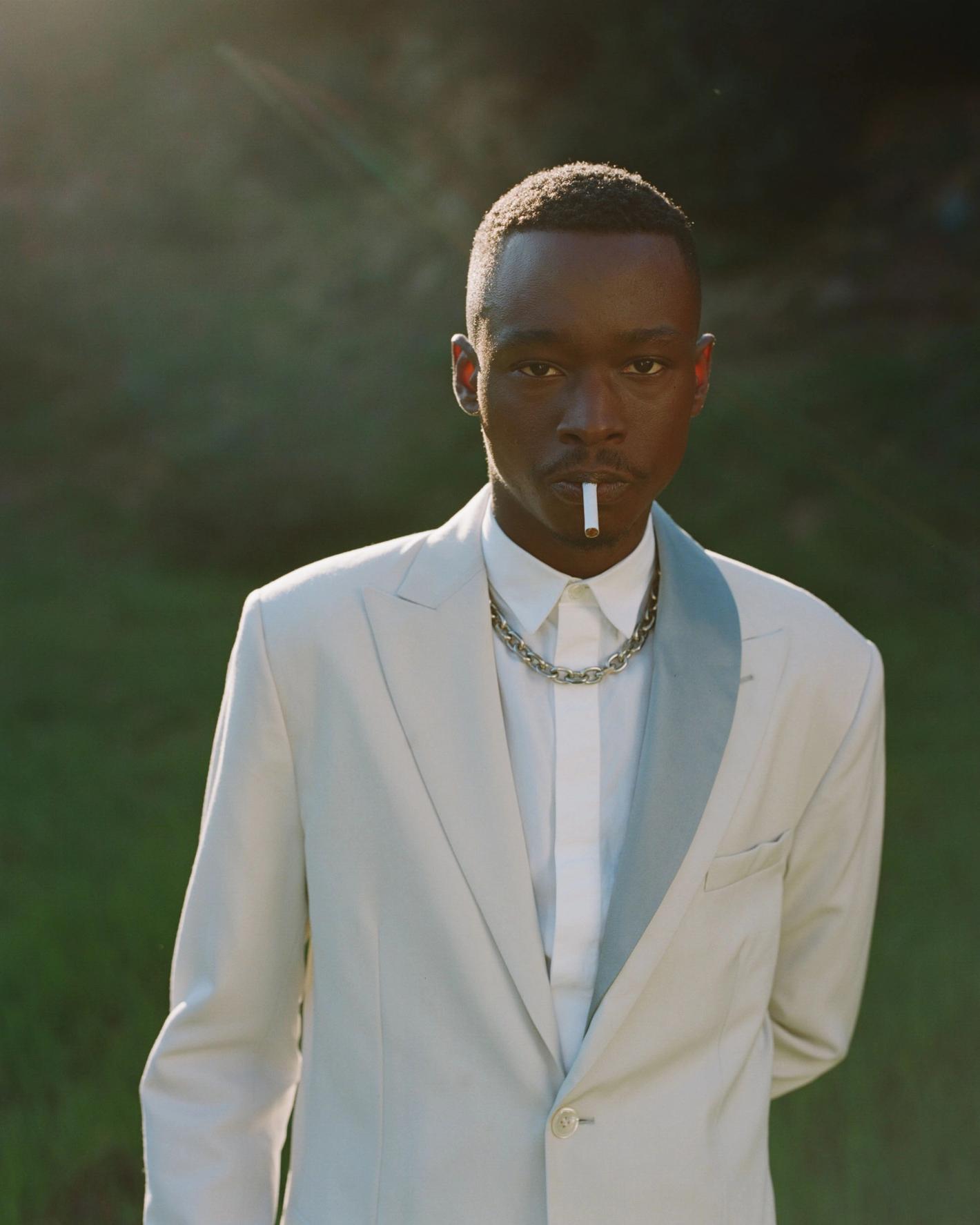 Close-up studio portrait of a Black male model in an off-white suit, buttoned shirt, chain necklace, and cigarette against a green background.