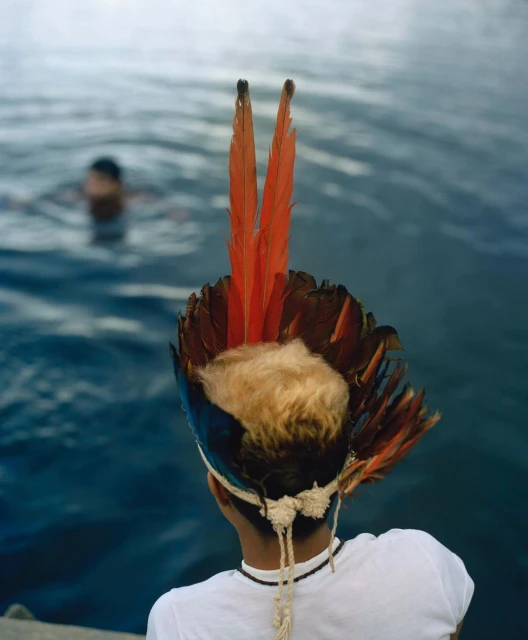 Person with feather headdress stands near water's edge as another person swims in the background on a cloudy day with natural lighting.