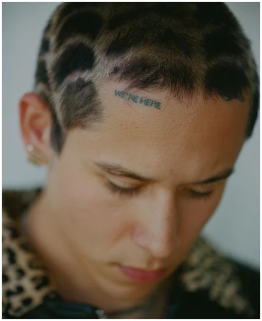 Close-up portrait of a young man with a short patterned haircut and a small forehead tattoo reading WE'RE HERE, photographed by Daniel Jack Lyons.