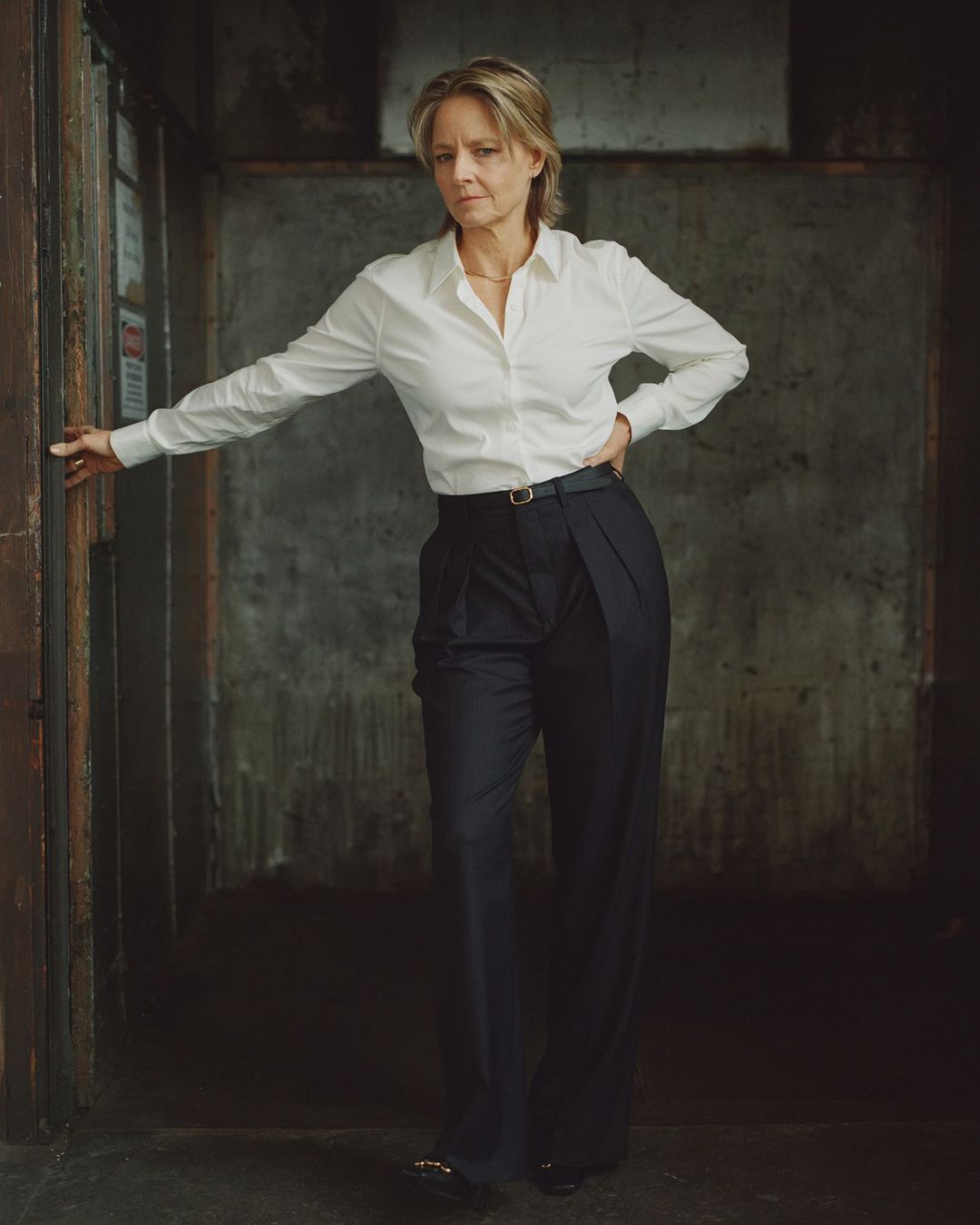 Indoor portrait of an adult woman in a white button-down shirt and dark slacks leaning on a door frame against a concrete wall.