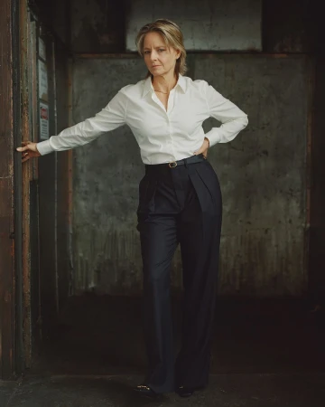 Indoor portrait of an adult woman in a white button-down shirt and dark slacks leaning on a door frame against a concrete wall.