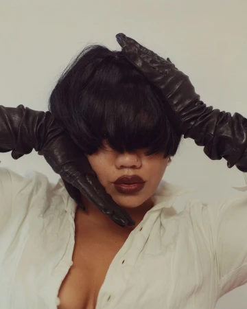 Portrait of a person wearing black leather gloves framing their face, styled with dark bob hair and a white collared shirt.