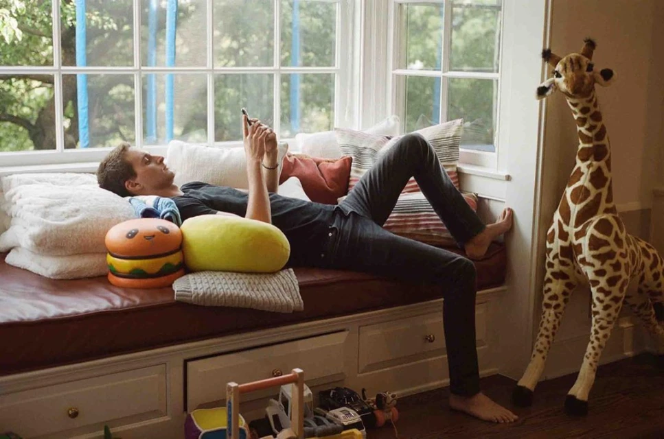 Adult male relaxes on a window seat with pillows, texting on a smartphone, with a large stuffed giraffe and kid's toys nearby.
