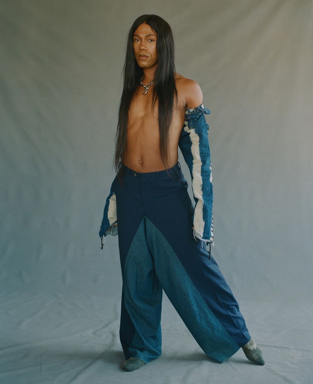 Person with long black hair poses in blue deconstructed-style wide-leg jeans and a top with torn, distressed sleeves in a studio.