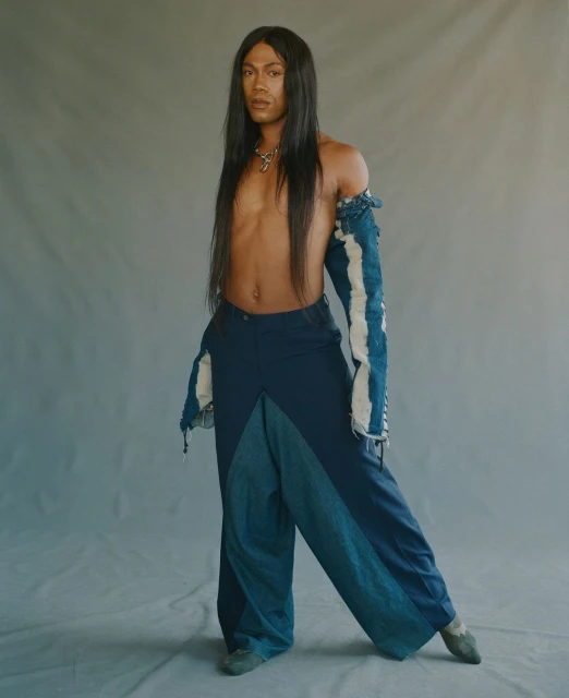 Person with long black hair poses in blue deconstructed-style wide-leg jeans and a top with torn, distressed sleeves in a studio.