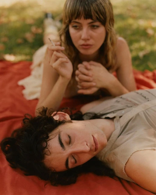 Two people relaxing on a red blanket outdoors in soft light. An adult female is holding a cigarette and a person is laying down with eyes closed.