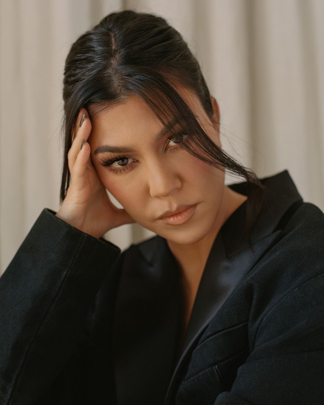 Close up portrait of Kourtney Kardashian wearing a black blazer with her hand resting against her head against a draped background.