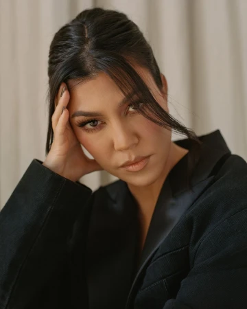 Close up portrait of Kourtney Kardashian wearing a black blazer with her hand resting against her head against a draped background.