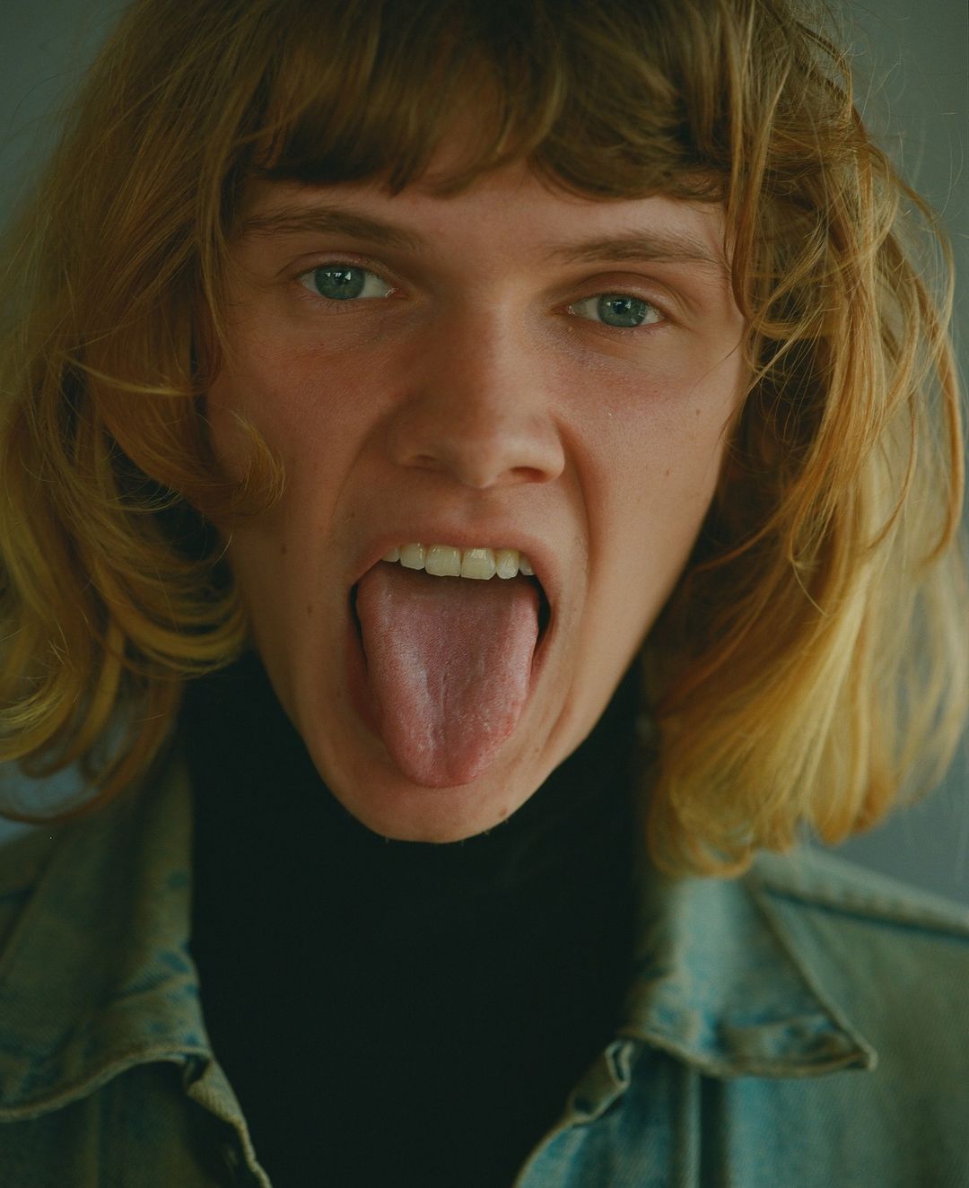 Close-up portrait of a young adult with blond hair and blue eyes, sticking their tongue out, wearing a denim jacket and black turtleneck.
