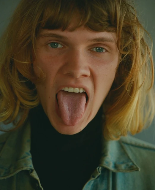 Close-up portrait of a young adult with blond hair and blue eyes, sticking their tongue out, wearing a denim jacket and black turtleneck.