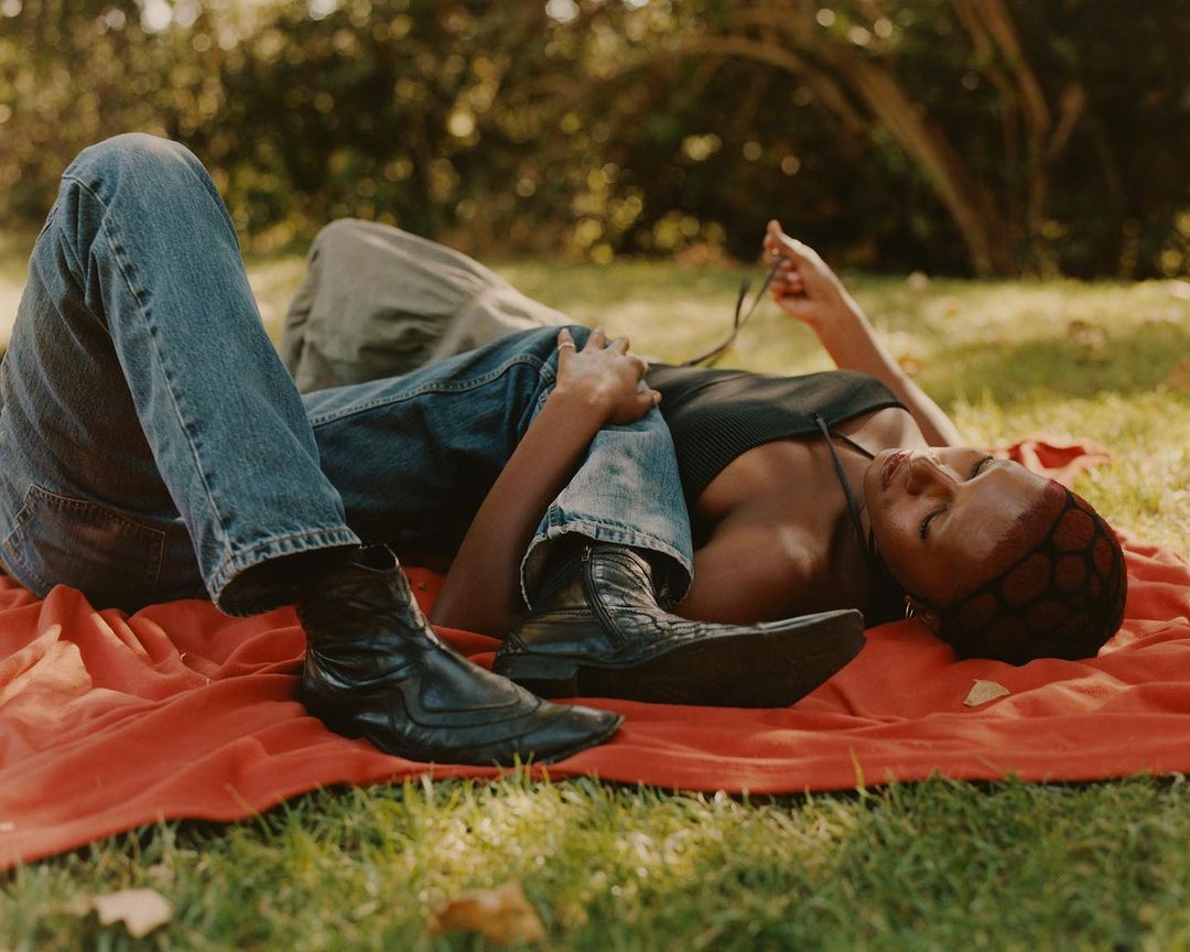 Two people, an adult woman and an adult, lie on a red blanket on a grass lawn in sunlight, wearing jeans and black boots.