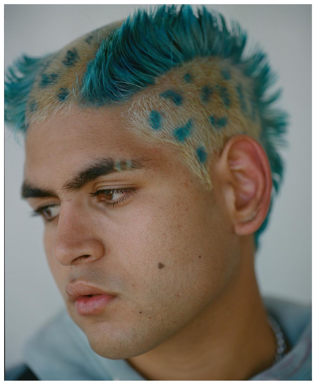 Close-up portrait of a young adult man with short blonde hair styled into a mohawk with turquoise dye and matching turquoise spots.