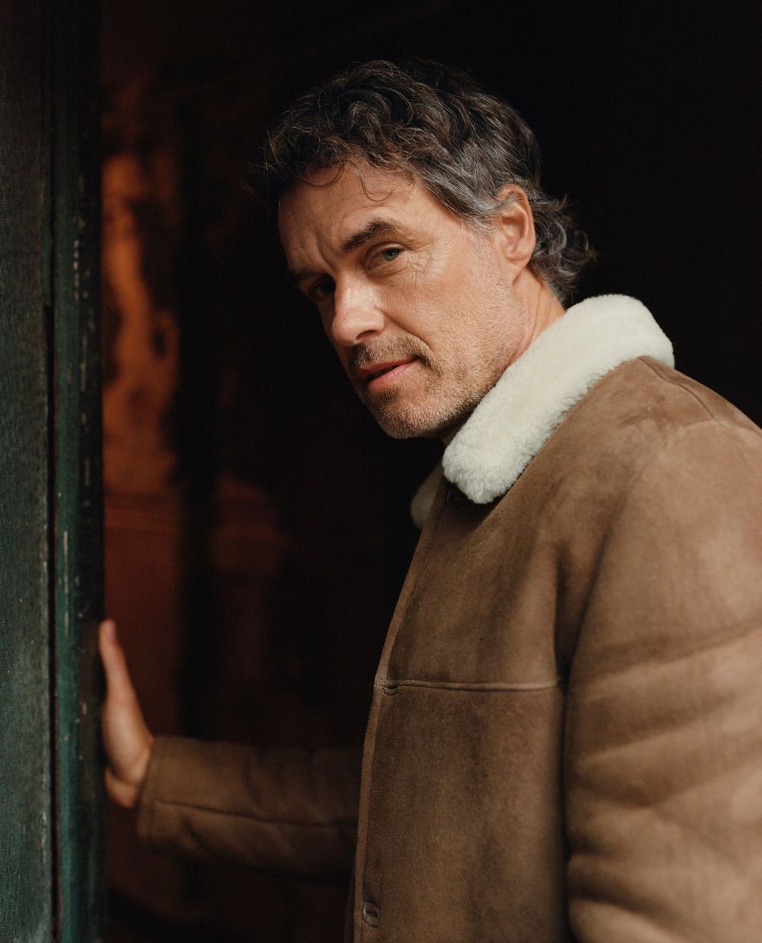 Portrait of a mature adult male with gray hair wearing a brown jacket with a white collar, looking toward the camera, dark background.