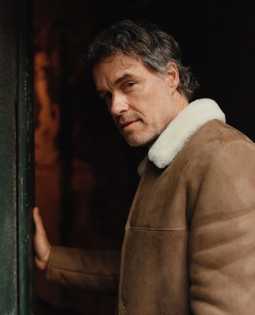 Portrait of a mature adult male with gray hair wearing a brown jacket with a white collar, looking toward the camera, dark background.