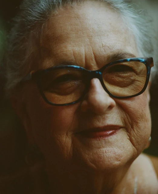 Close-up portrait of an elderly woman with grey hair and glasses, conveying warmth with a slight smile in soft, natural light.