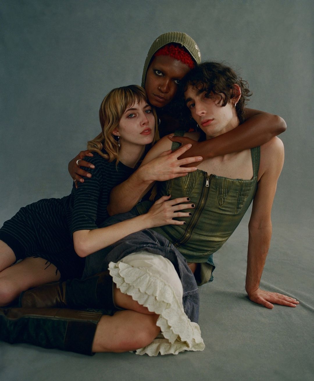 Three people posing indoors: a woman in a dress, a person in a corset, and a person in a hoodie, all embracing in a studio setting.