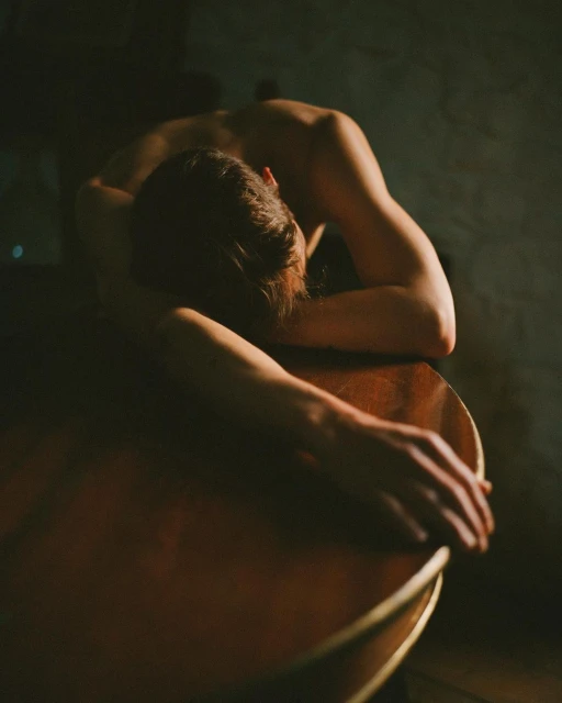 Artistic photo of a bare-chested adult resting on a wooden guitar, captured in dim, moody lighting inside an intimate indoor setting.