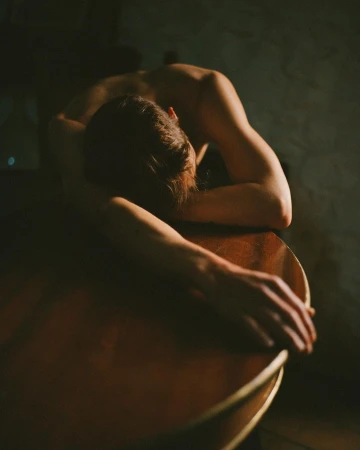 Artistic photo of a bare-chested adult resting on a wooden guitar, captured in dim, moody lighting inside an intimate indoor setting.