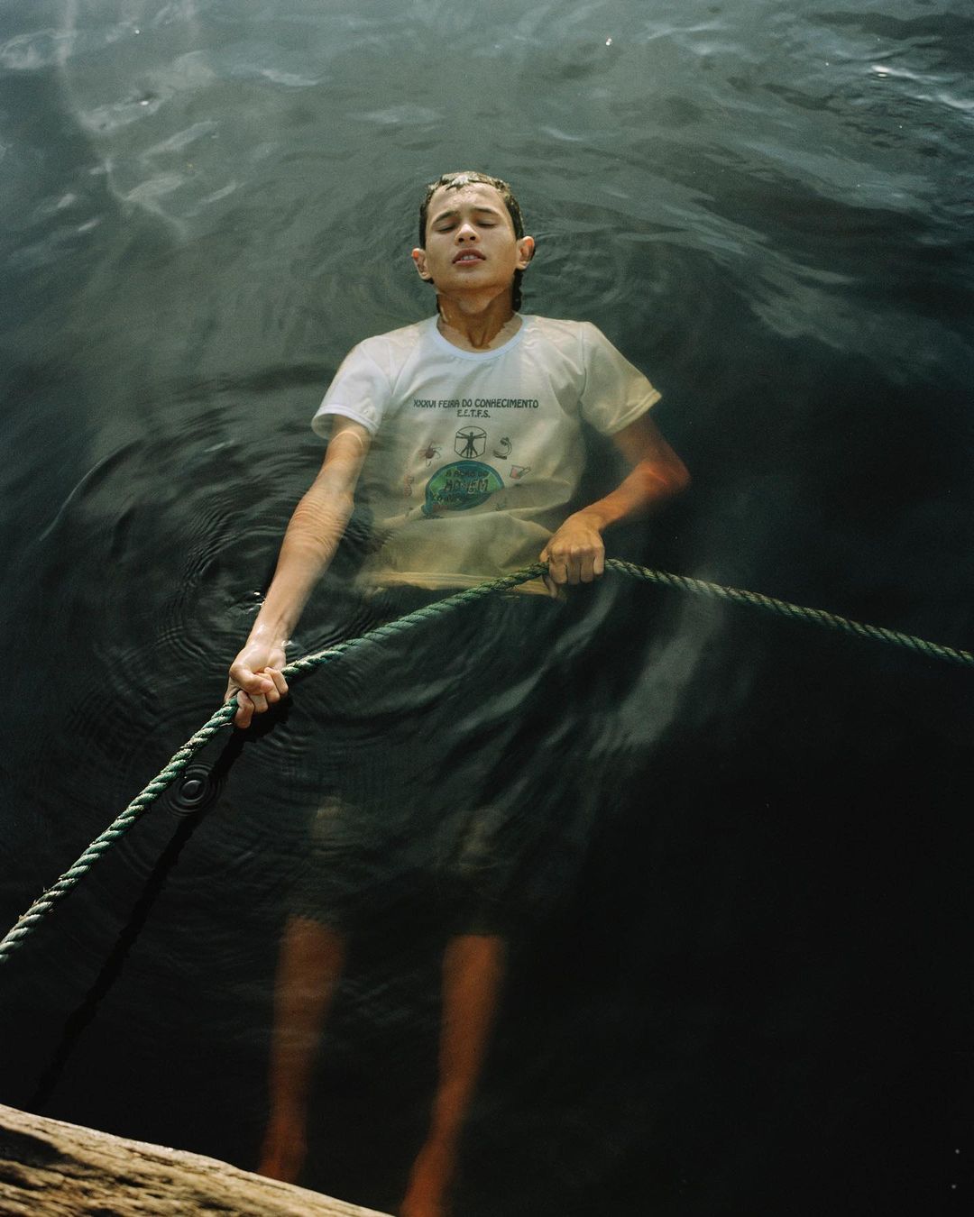 A child stands chest-deep in dark water, gripping a rope, eyes closed, wearing a white t-shirt, creating a somber, reflective portrait.