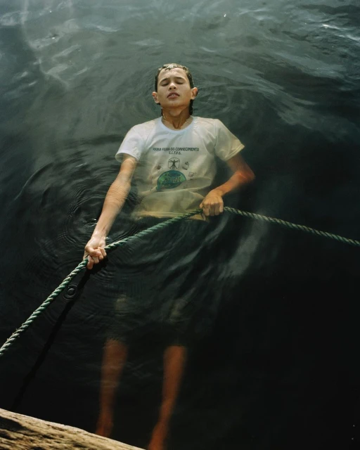 A child stands chest-deep in dark water, gripping a rope, eyes closed, wearing a white t-shirt, creating a somber, reflective portrait.