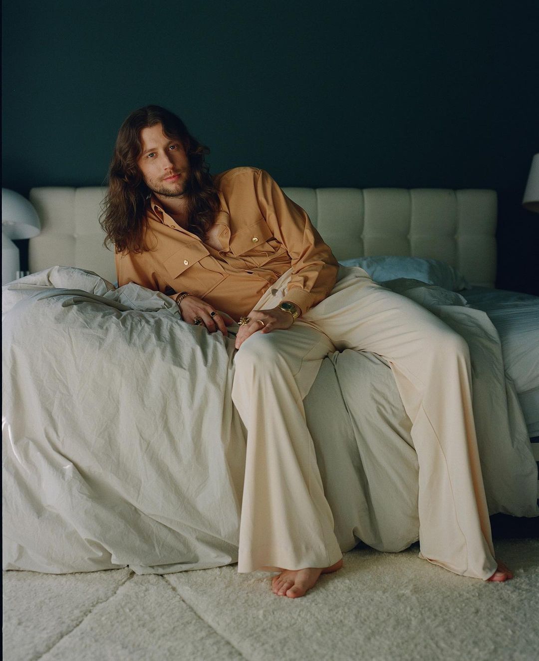 A fair-skinned man with long brown hair in an orange shirt and cream pants reclines on a bed with a blue wall in the background.